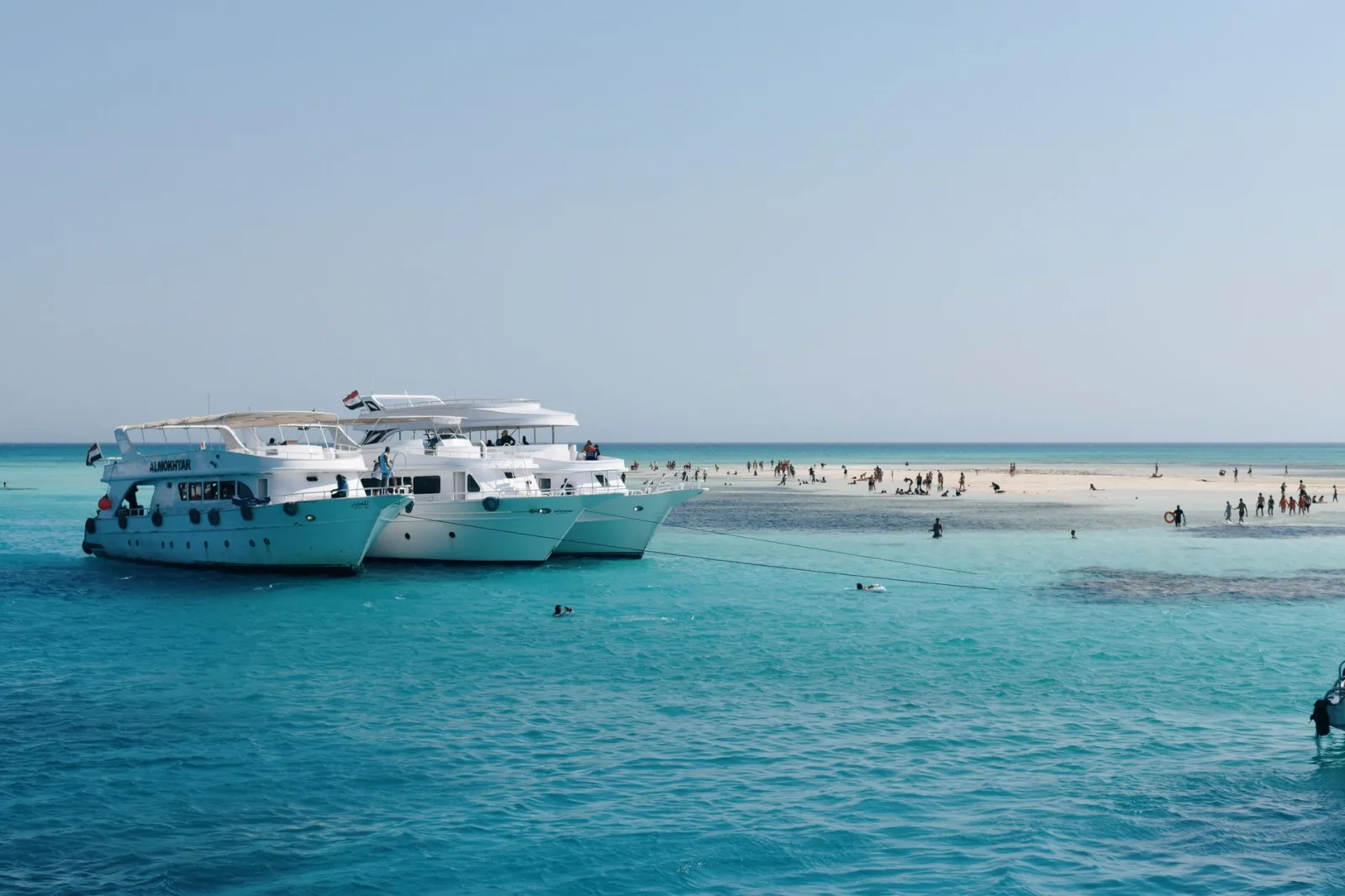 Yachts anchored near a sandy island with people enjoying a sunny day.