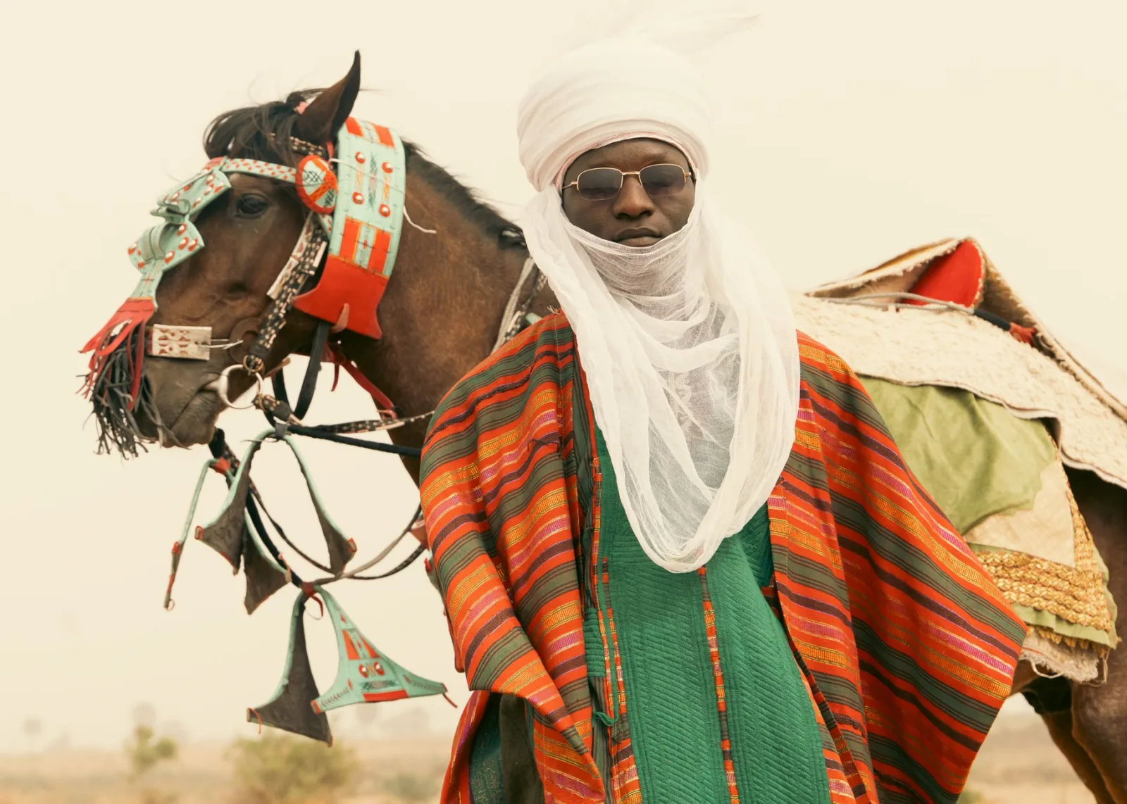 Person in traditional colorful attire stands beside a decorated horse in a desert setting, exuding cultural richness.