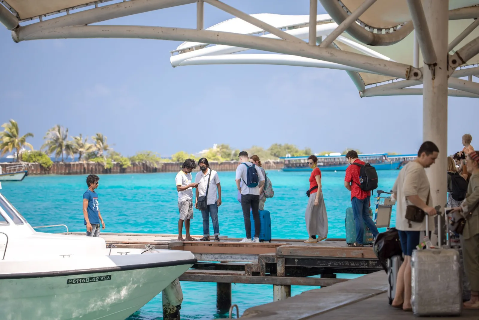 People with luggage waiting on a dock with turquoise ocean and clear sky in a tropical setting.