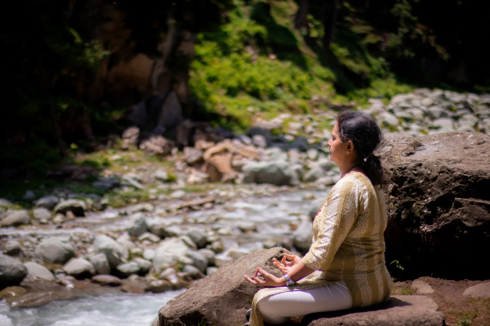 Woman practicing meditation near a tranquil river in a rocky, lush environment.