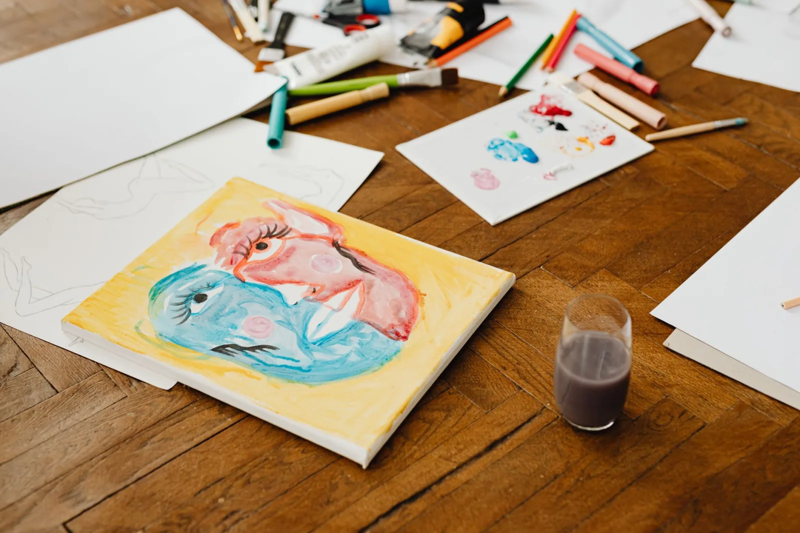 Art supplies scattered on a wooden floor, featuring vibrant paintings and tools for creative projects.