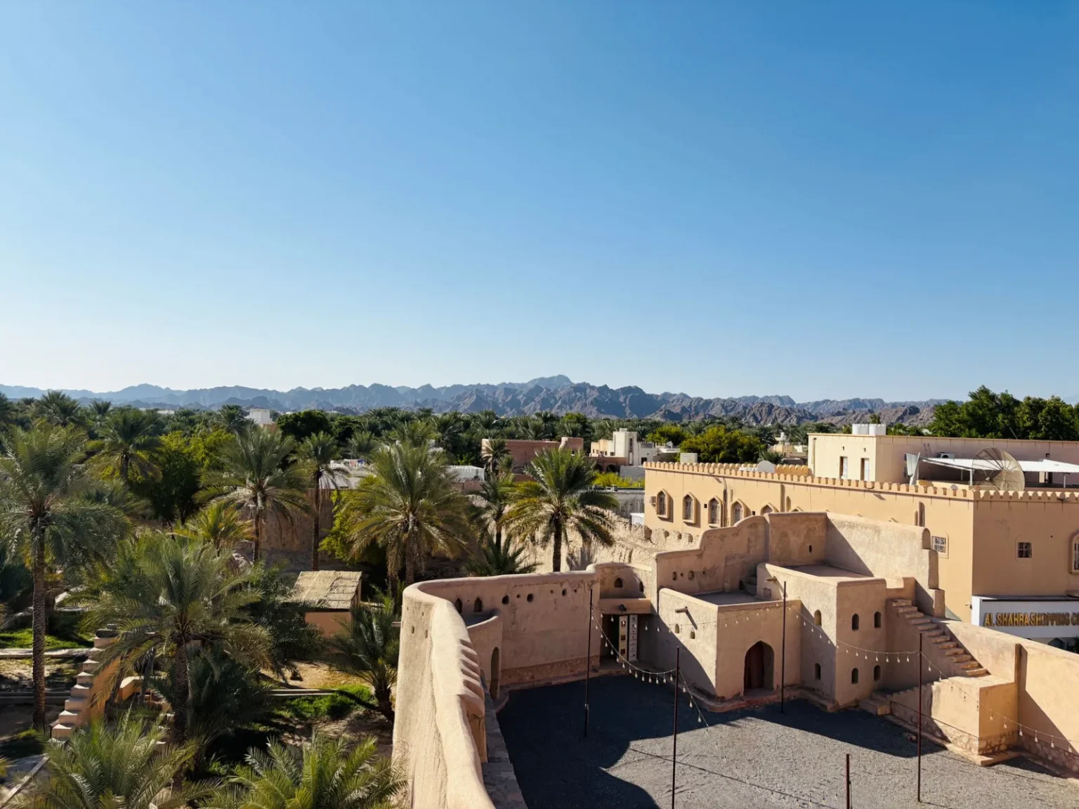 View of Nizwa Fort and the surrounding oasis against Hajar Mountains under a clear blue sky.