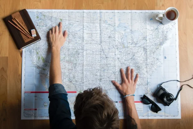 Top view of anonymous young male in casual clothes sitting on wooden floor with photo camera and coffee cup and pointing on paper map while planning trip route