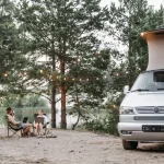 A couple enjoys a serene camping experience by the lake with a camper van and string lights.