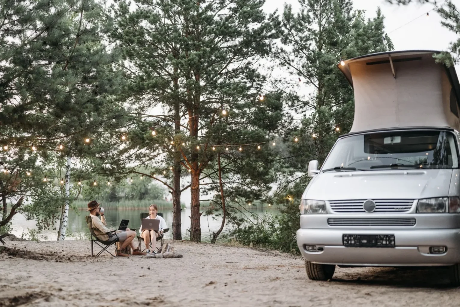 A couple enjoys a serene camping experience by the lake with a camper van and string lights.