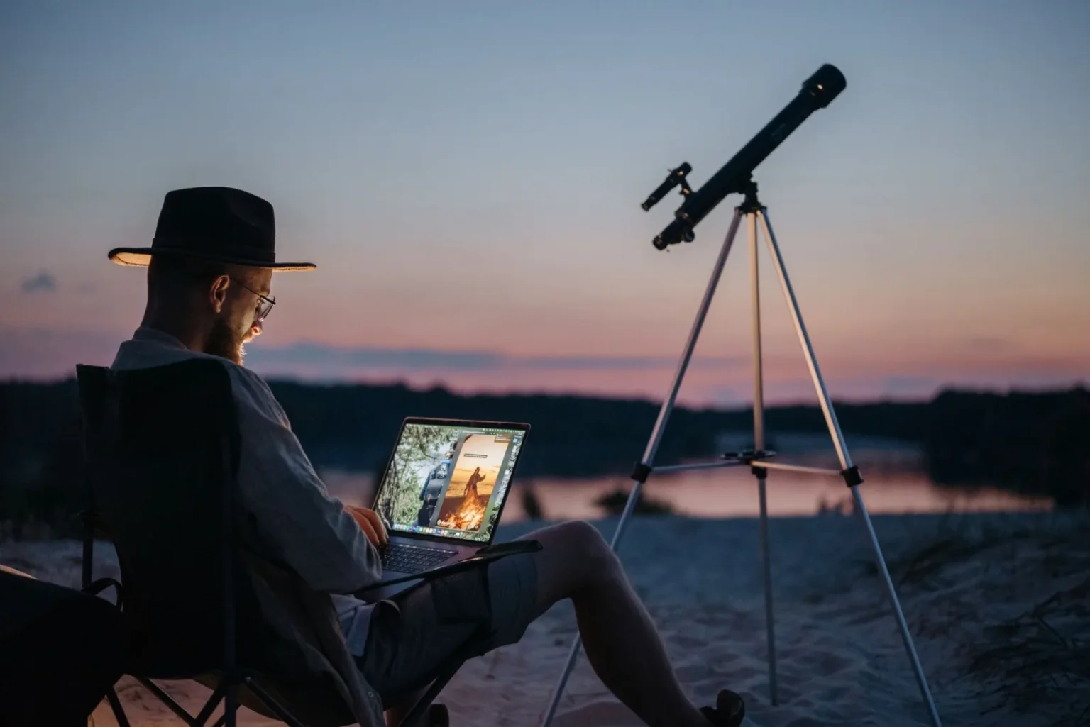 A man enjoys a camping trip by the lake using a laptop and telescope at sunset.