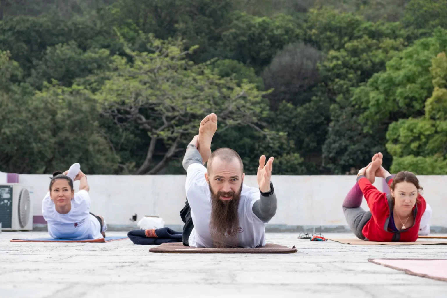 A diverse group of adults practicing yoga on a rooftop in Rishikesh, surrounded by greenery.