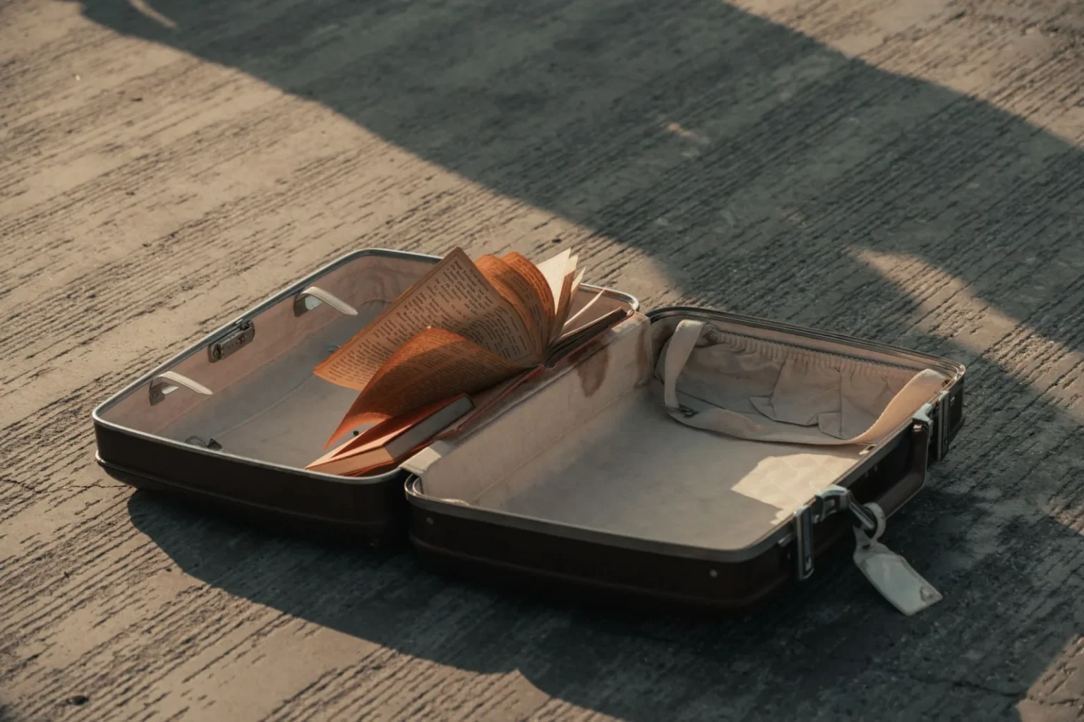 An open suitcase with a book lying on a concrete surface, casting a shadow.