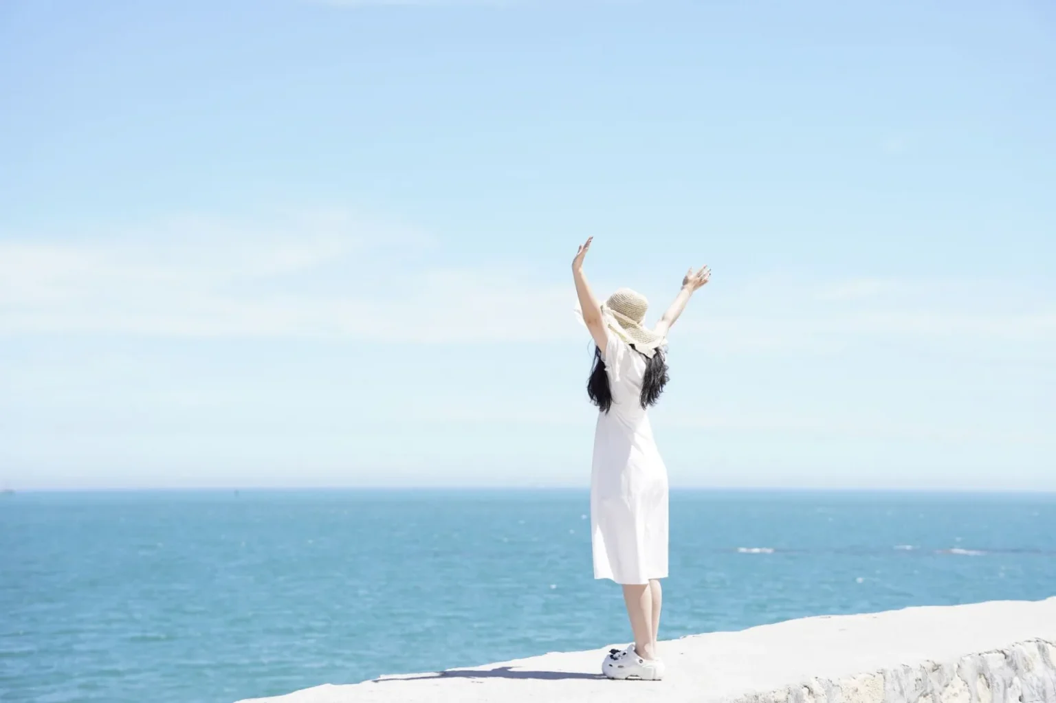 Woman in a white dress and hat raises arms joyfully by the ocean on a sunny day.