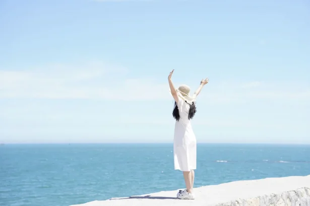Woman in a white dress and hat raises arms joyfully by the ocean on a sunny day.