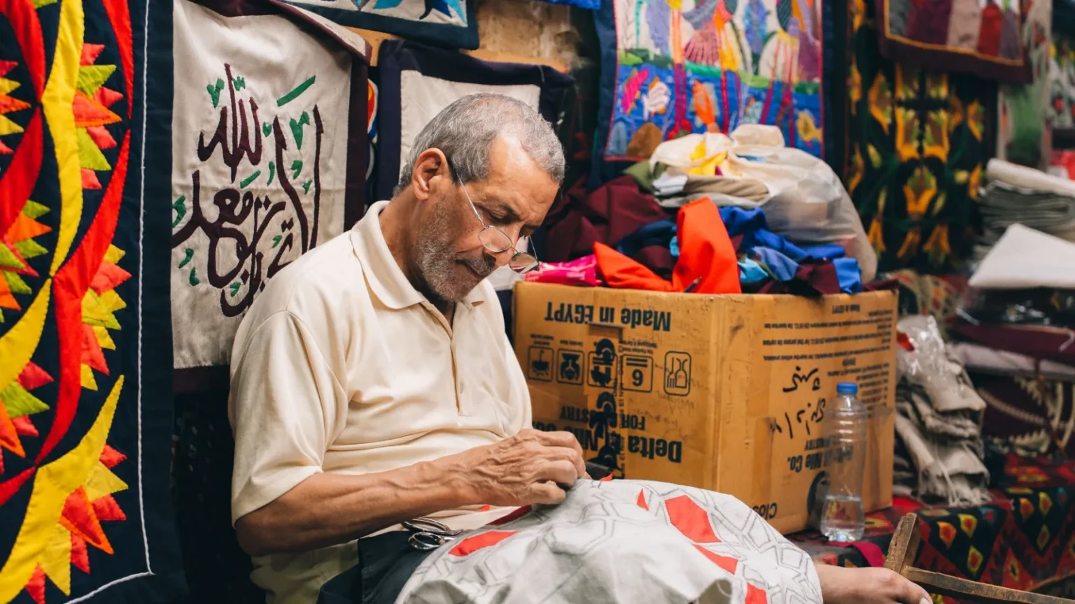 Elderly male artisan sewing traditional Egyptian textiles in a colorful workshop.