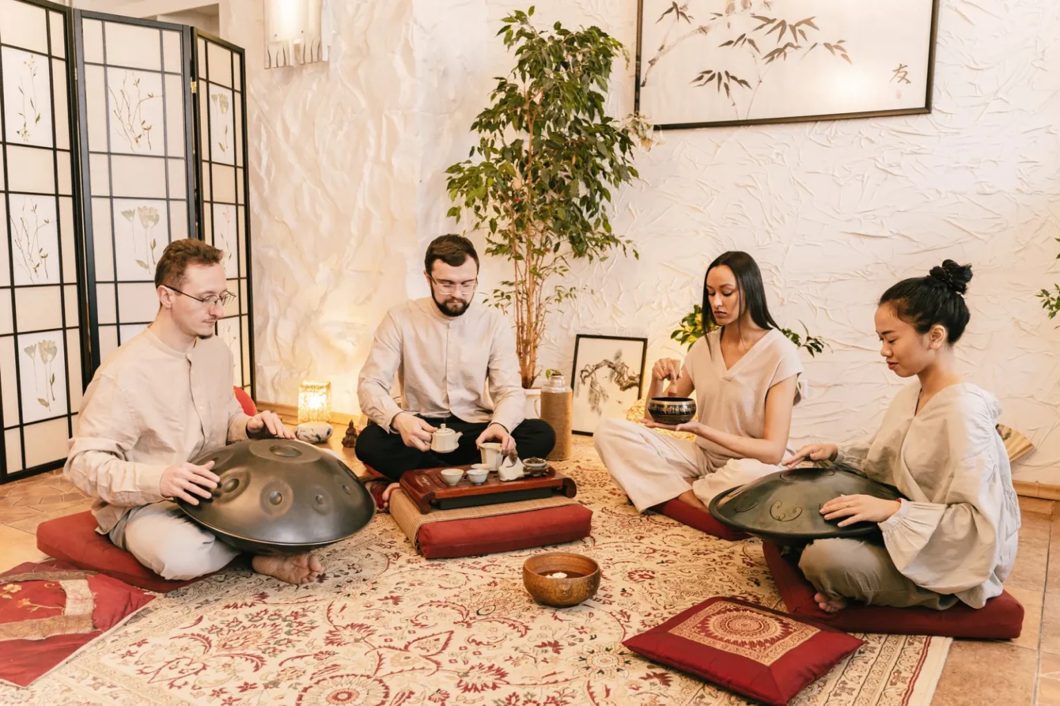A diverse group meditates with handpans and tea in a serene, indoor setting.