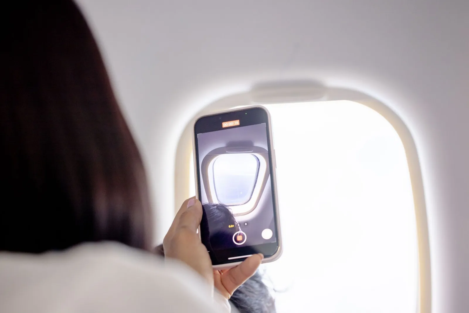 A woman records a video of the sky through an airplane window using her smartphone.