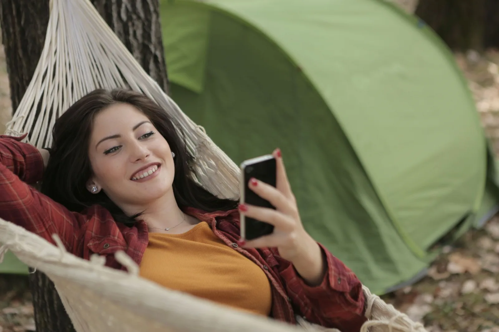Smiling woman lying in a hammock outdoors, enjoying a sunny day and using her smartphone.