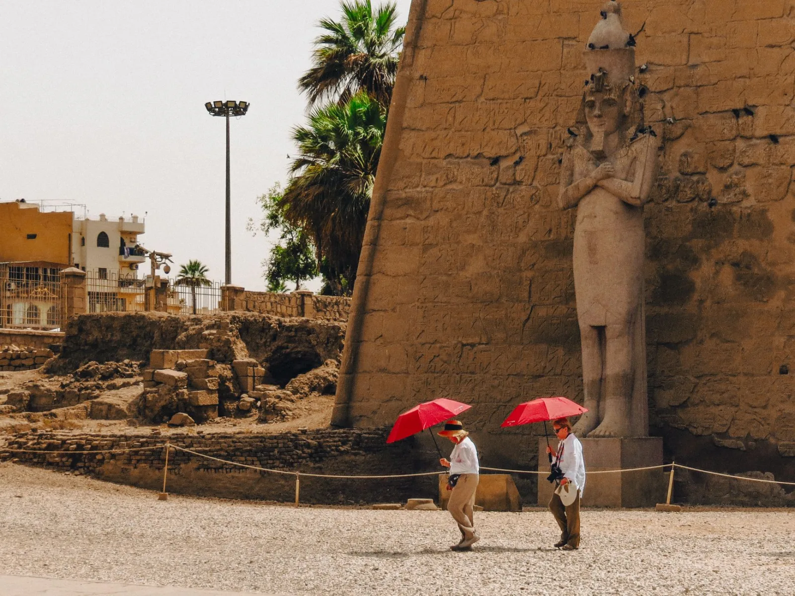 Two tourists walk past an ancient statue at Luxor Temple, Egypt, carrying red umbrellas.