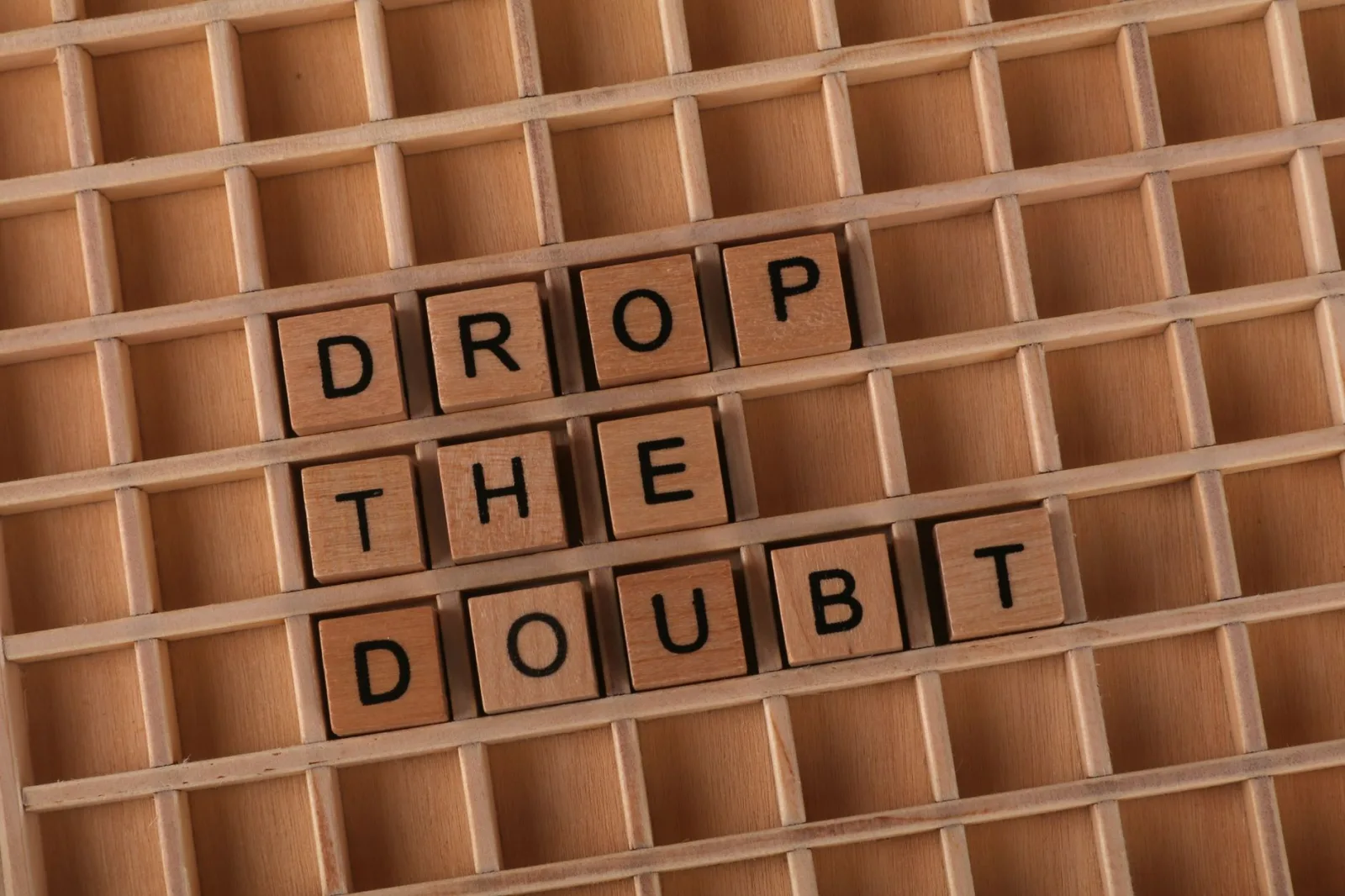 Wooden letter blocks arranged to form 'DROP THE DOUBT' on wooden grid background.