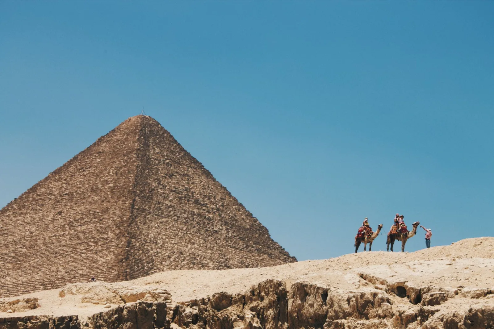 Camels and riders traverse the desert near the Great Pyramid of Giza under a blue sky.