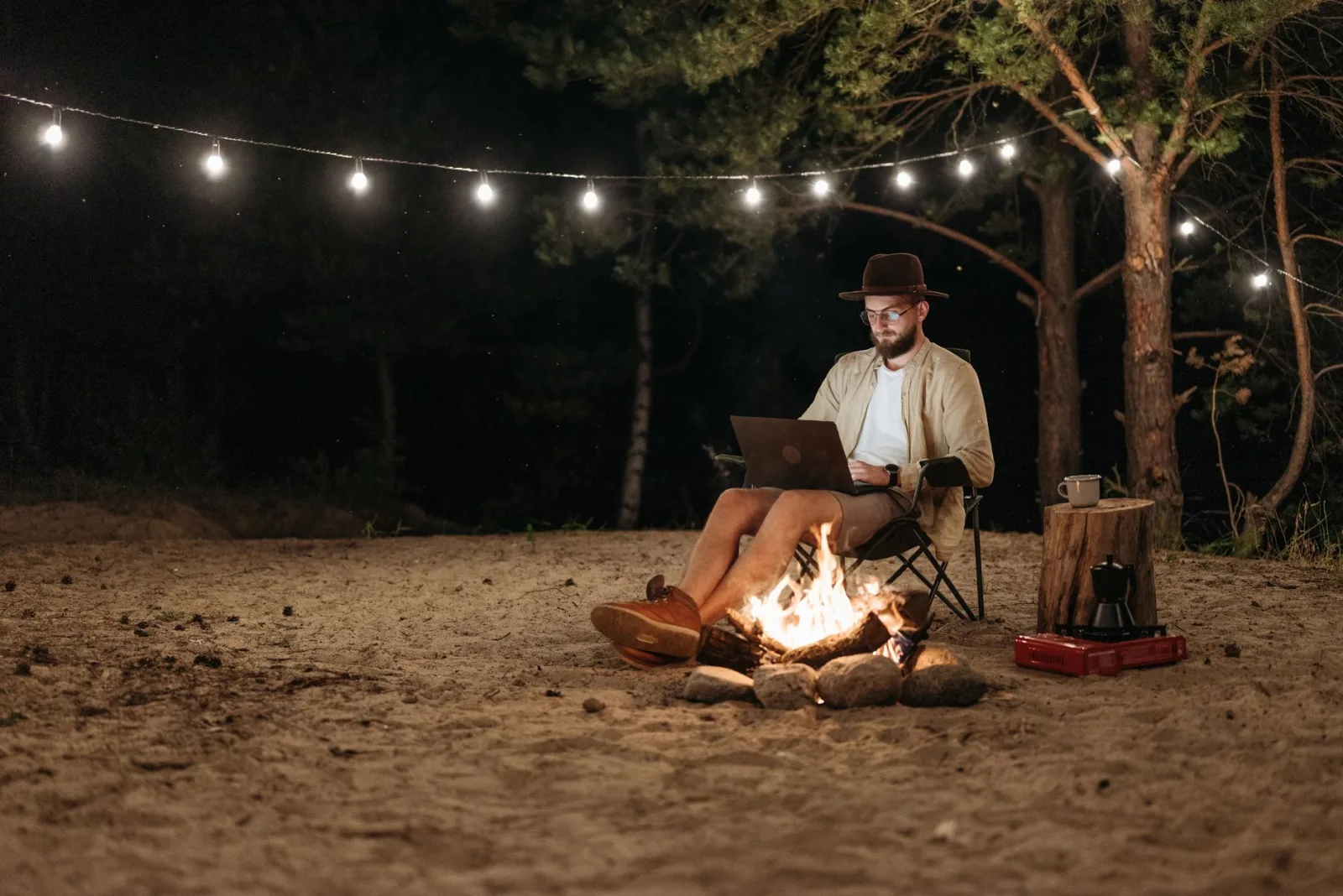 Digital nomad working remotely by a campfire with a laptop under string lights in the forest.