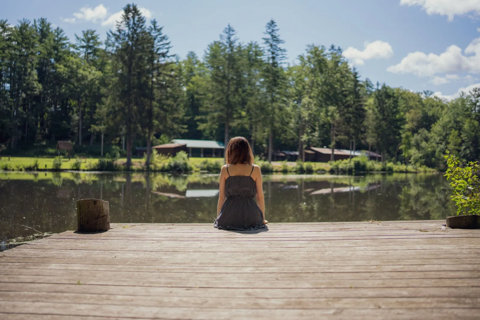 Serene view of a woman sitting on a wooden dock by a peaceful lakeside with lush trees in the countryside.
