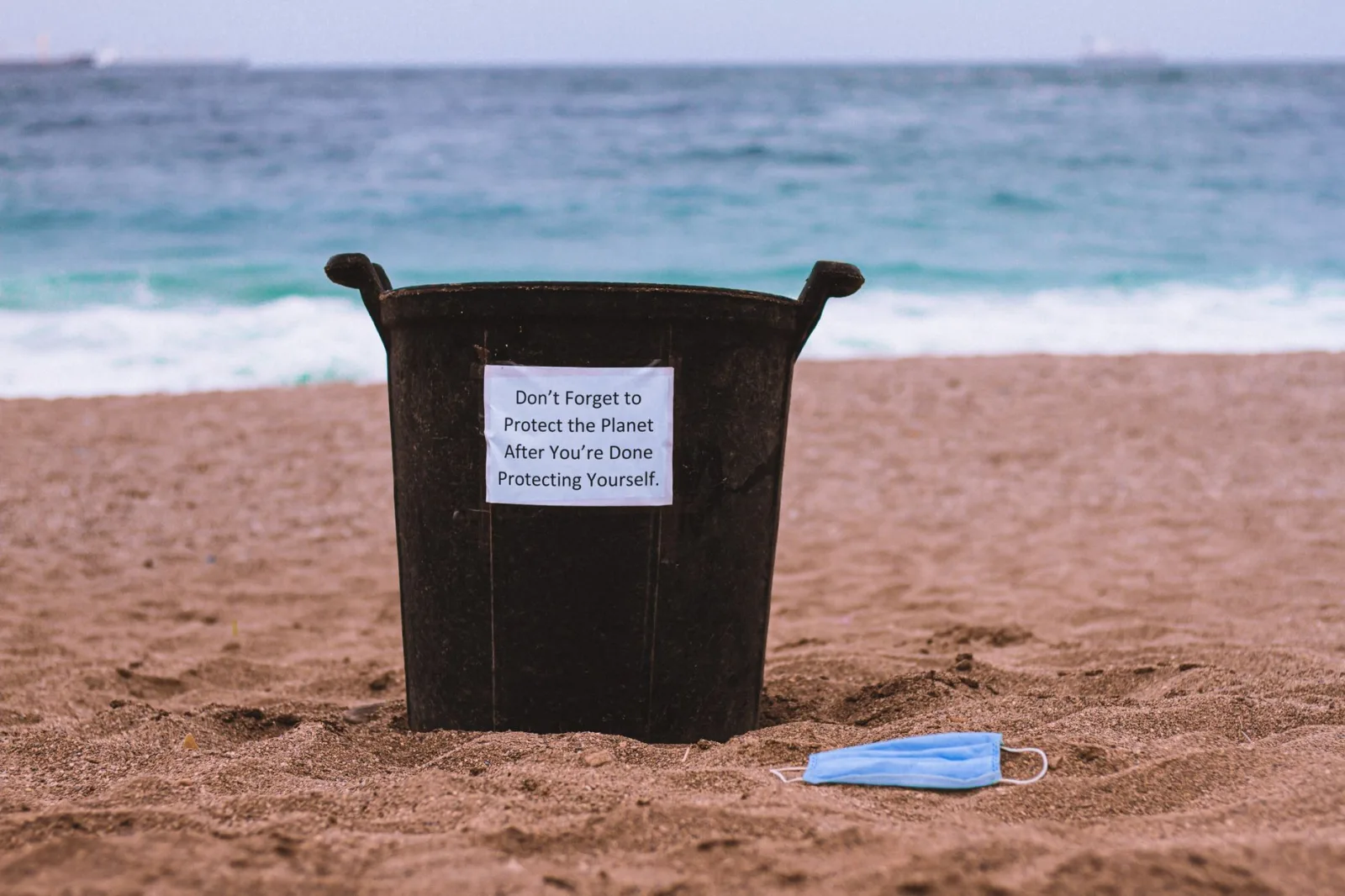A trash bin on the beach highlights environmental protection sustainability with a discarded mask.