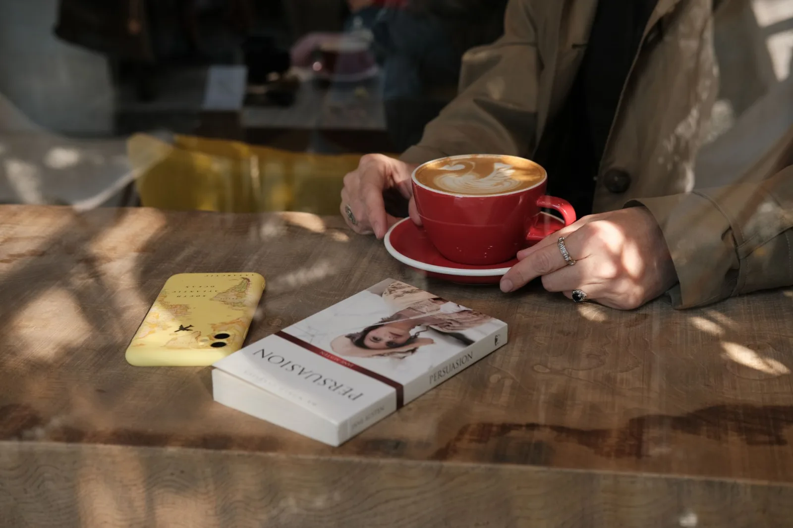 Person enjoying coffee with a book at a sunlit cafe table, creating a cozy atmosphere.