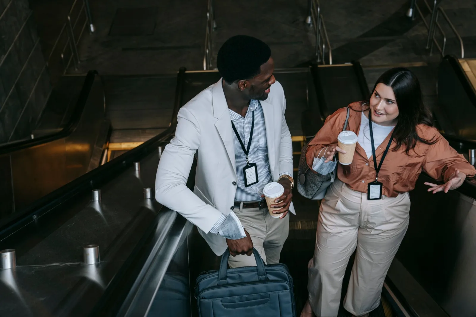 Two colleagues conversing with coffee cups on an indoor escalator.