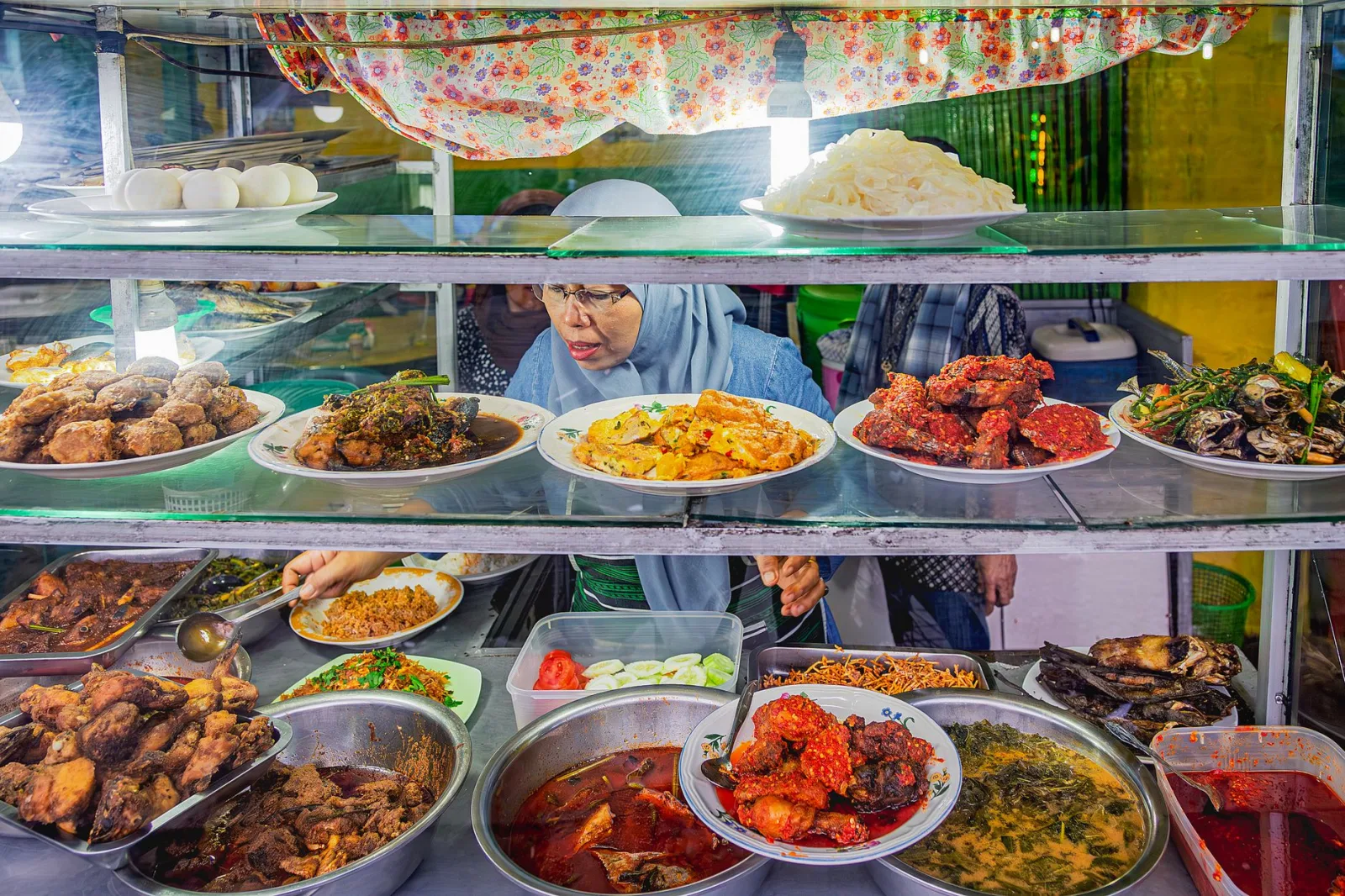 Colorful Indonesian street food stall with a variety of traditional dishes and a vendor serving customers.