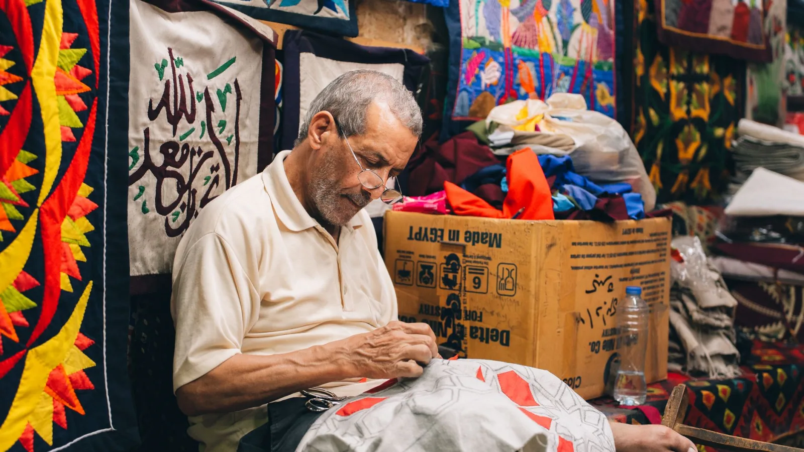 Elderly male artisan sewing traditional Egyptian textiles in a colorful workshop.