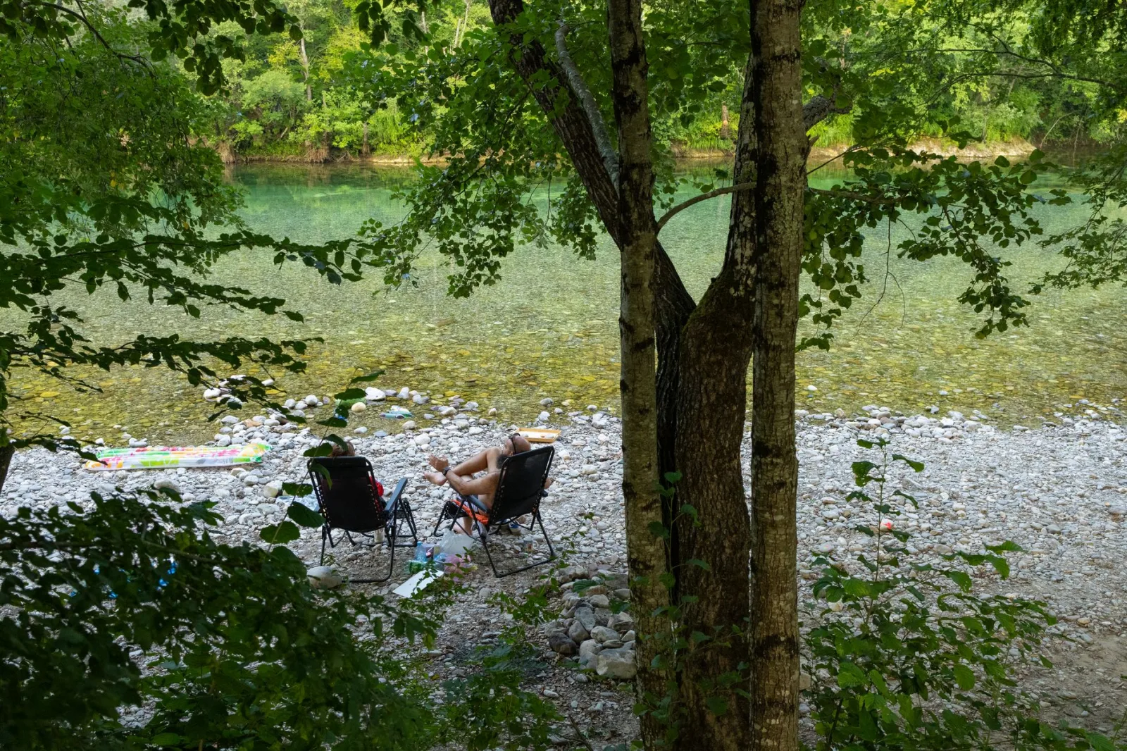 Two people relaxing on chairs by a clear riverbank surrounded by lush greenery.