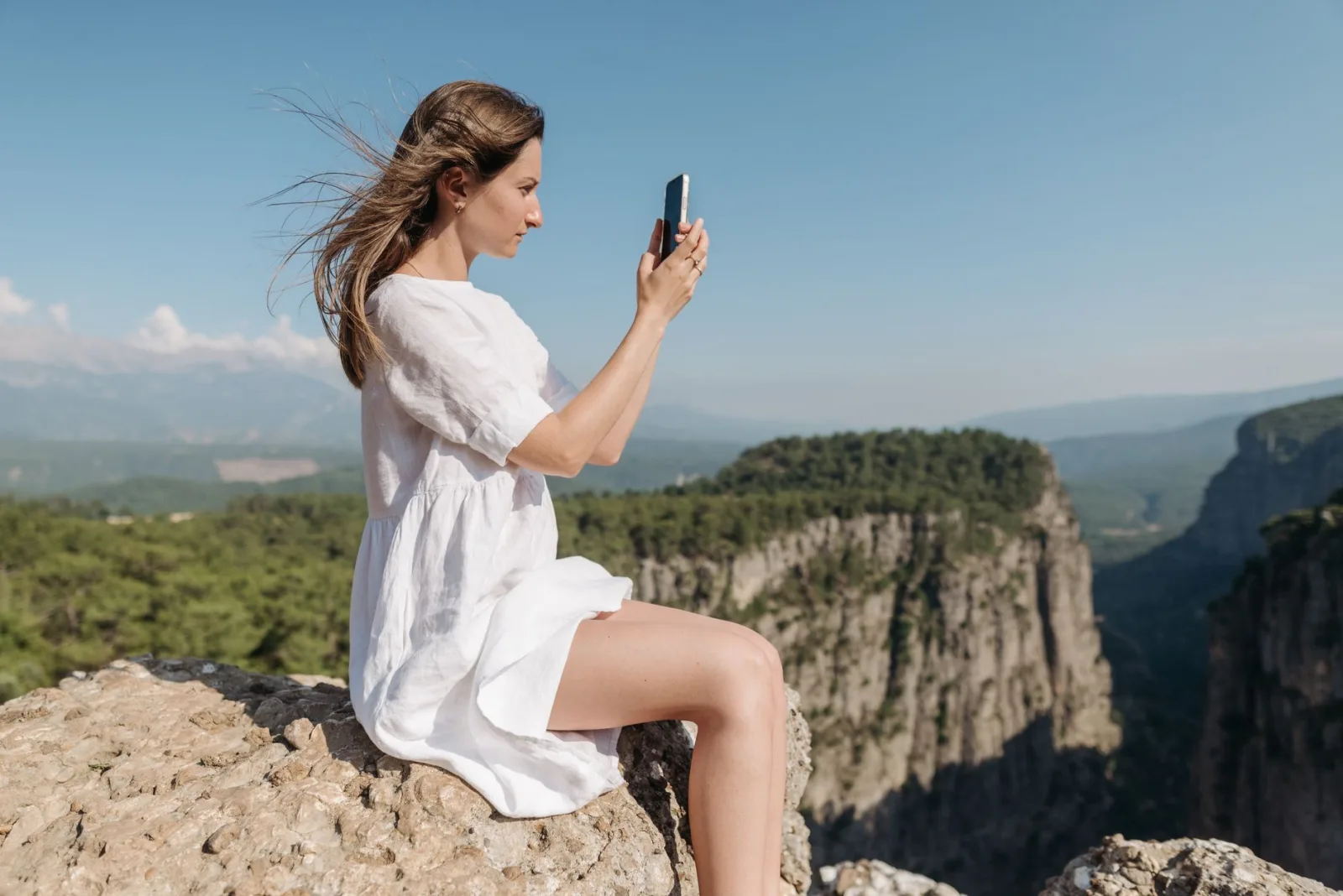 A woman in a white dress sits on a rock capturing a stunning mountain view.