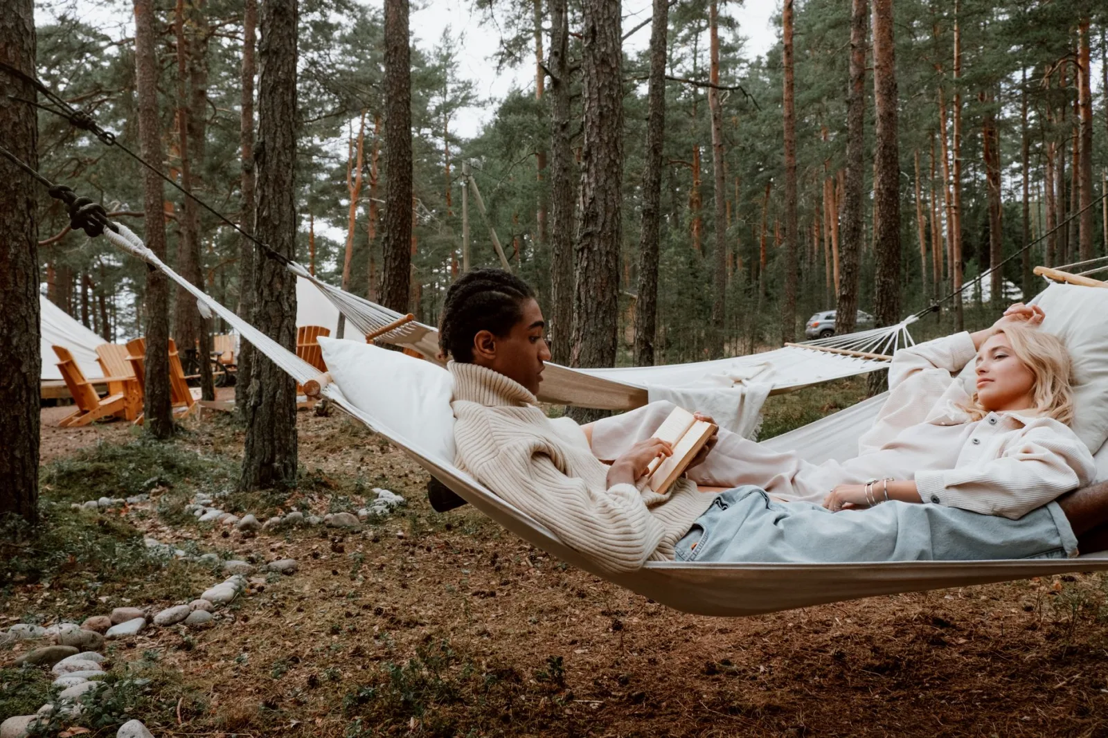 A man reads and a woman rests in a hammock amidst a serene forest setting, enjoying nature.