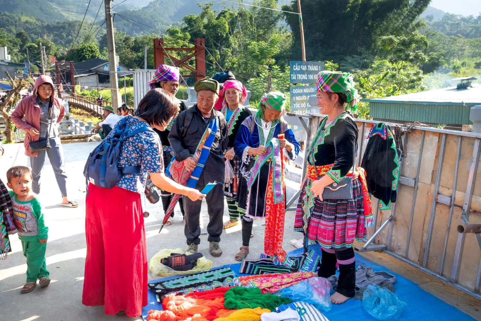 Smiling Asian female seller talking to anonymous buyer on pavement with ornamental fabrics in local market