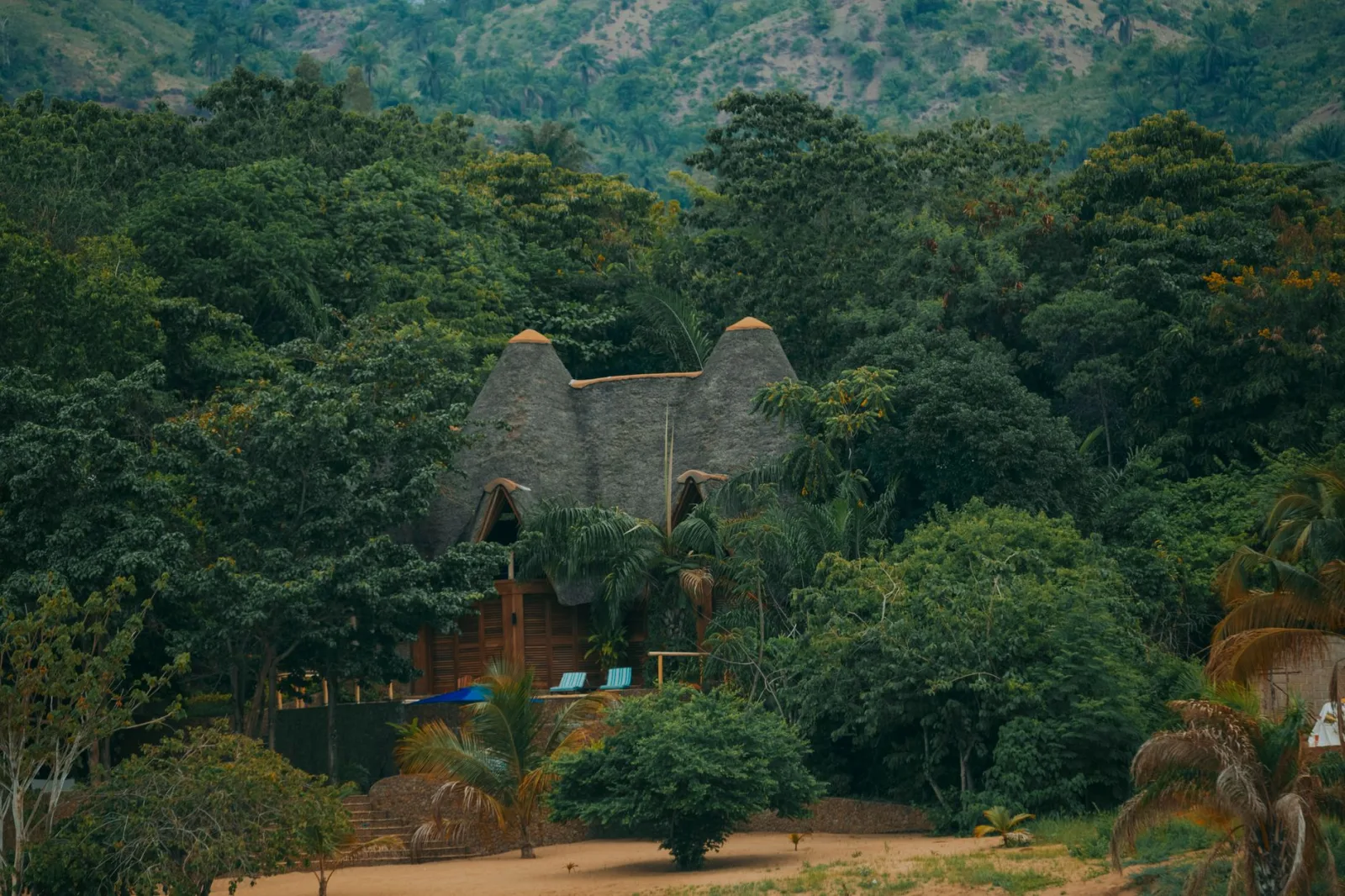 A rustic lodge with a thatched roof surrounded by lush greenery and hills.