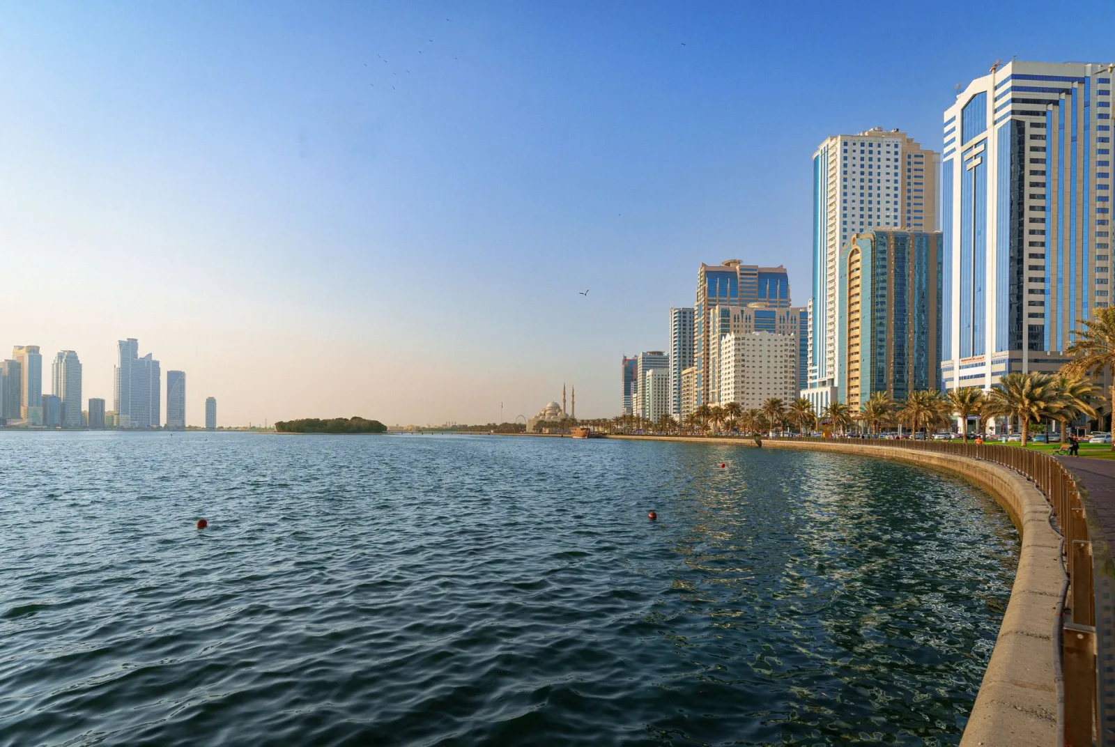 Beautiful daytime view of Sharjah's waterfront skyline with modern skyscrapers and calm blue waters.