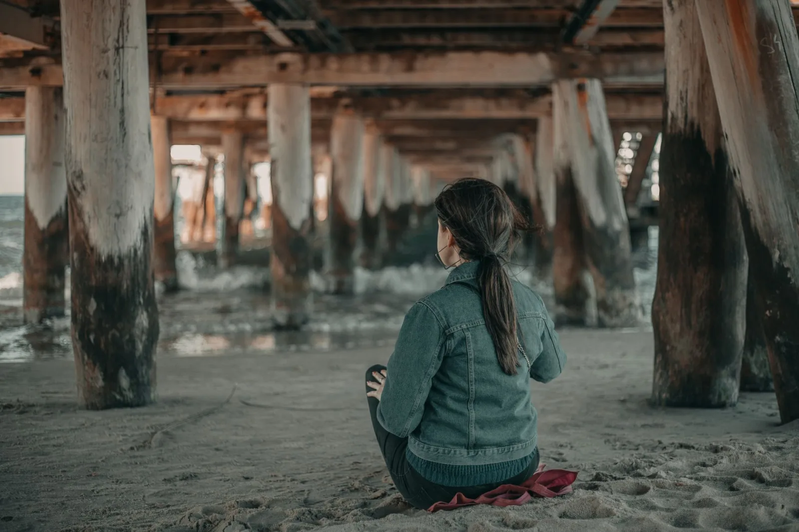 Back view of anonymous female tourist sitting on sandy coast near rippling sea under shabby wooden bridge in coastal terrain