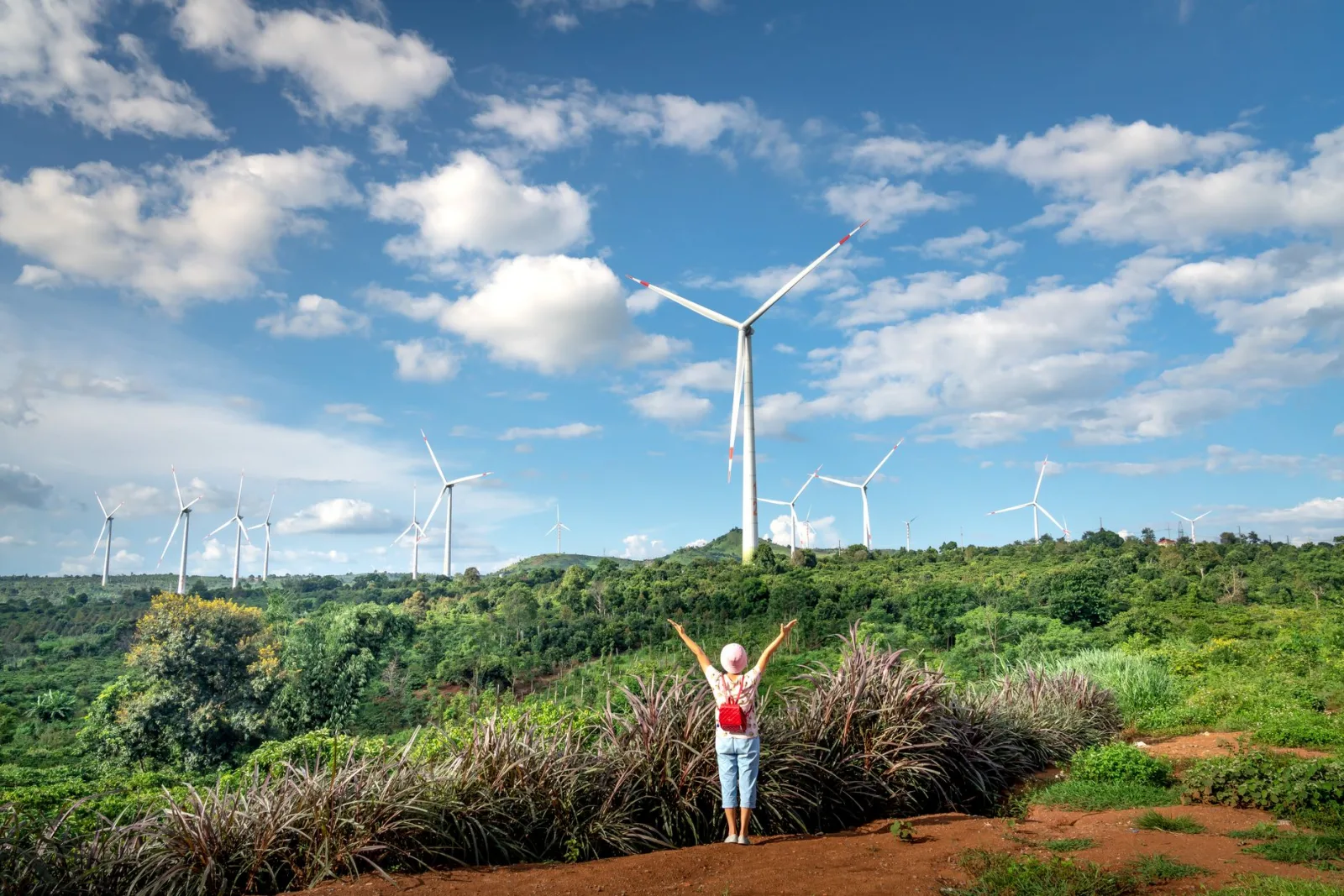 A woman joyfully raises her arms in front of wind turbines on a lush green hillside under a blue sky.
