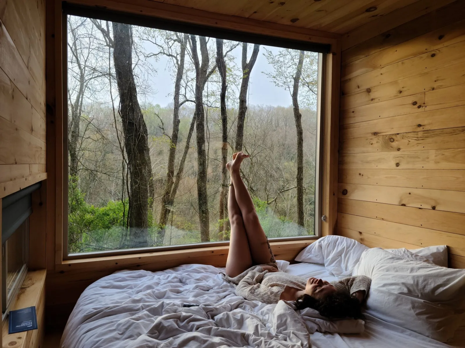 Woman relaxing on a bed in a cozy cabin, enjoying the view of the forest through a large window.