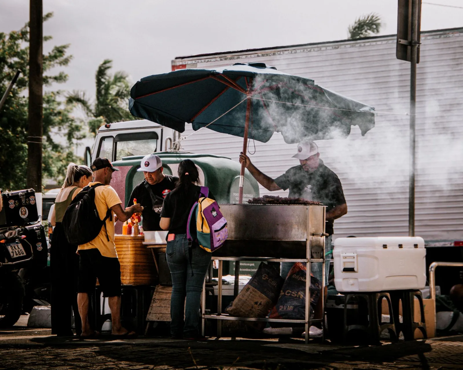 Street food vendor in action under umbrella serving customers. Smoky atmosphere adds charm.