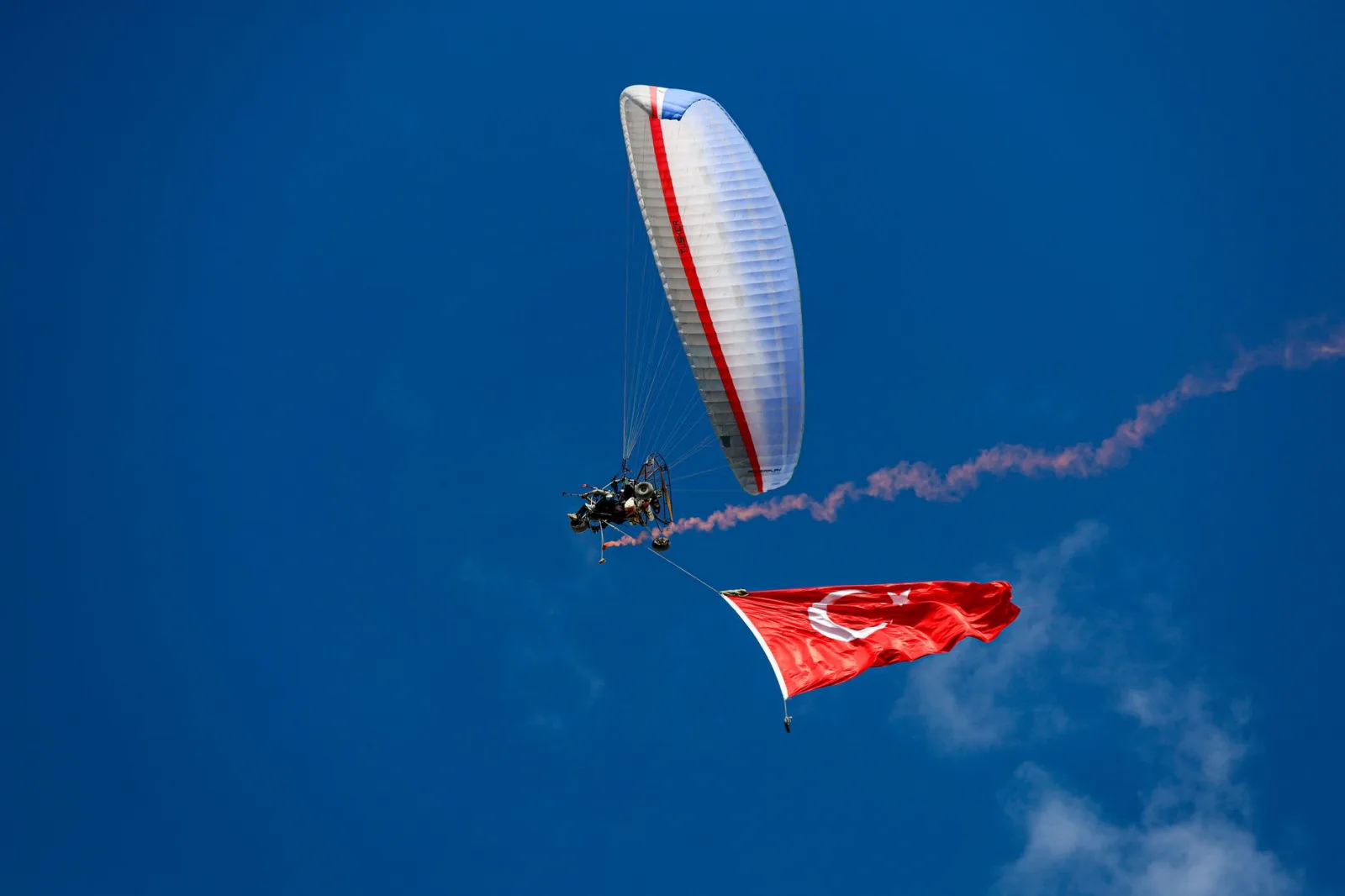 A paraglider flying high in a clear blue sky, adorned with the Turkish flag below.