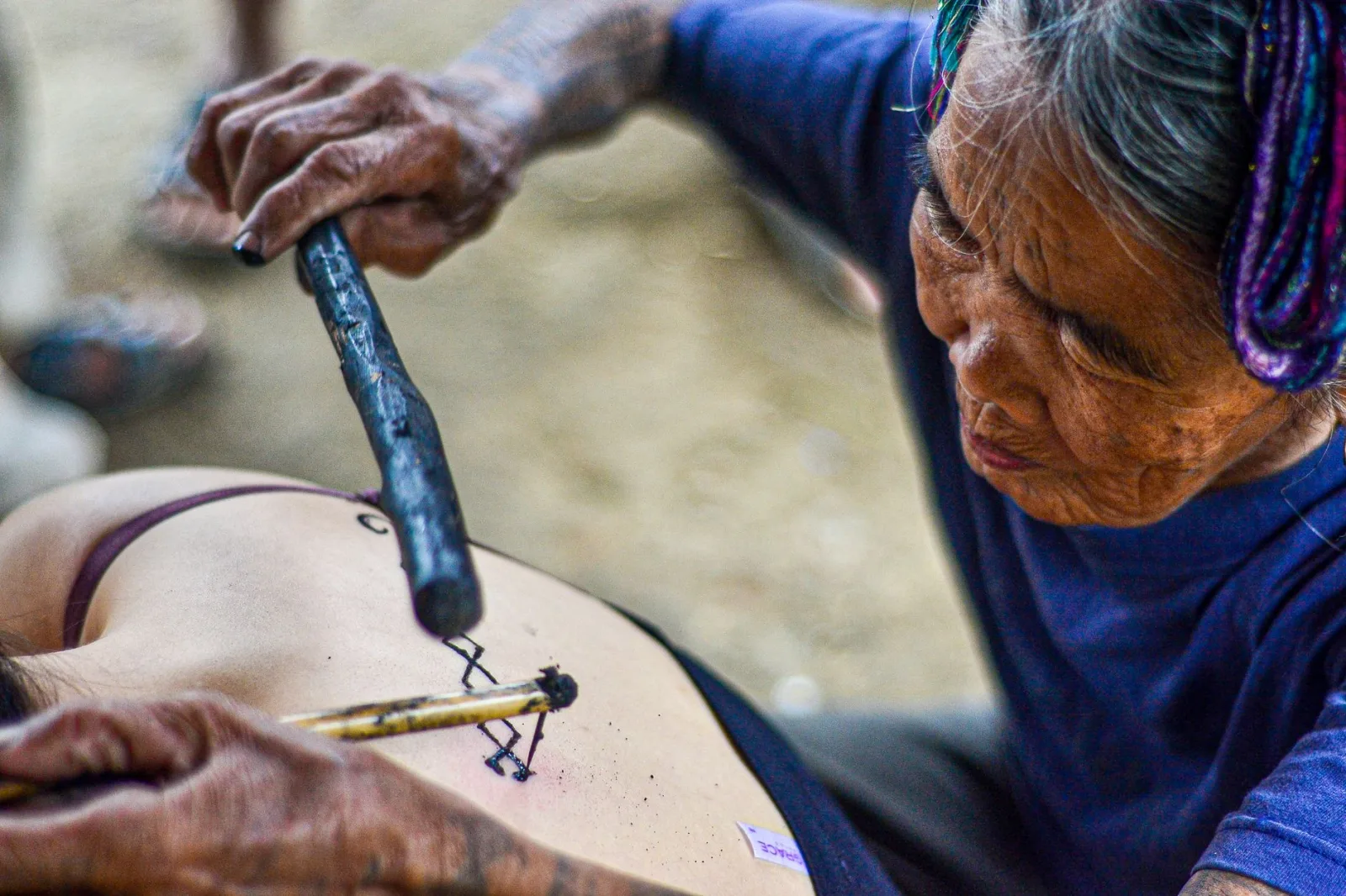 Elderly woman performing traditional hand-tap tattooing on a younger person's back in Kalinga, Philippines.
