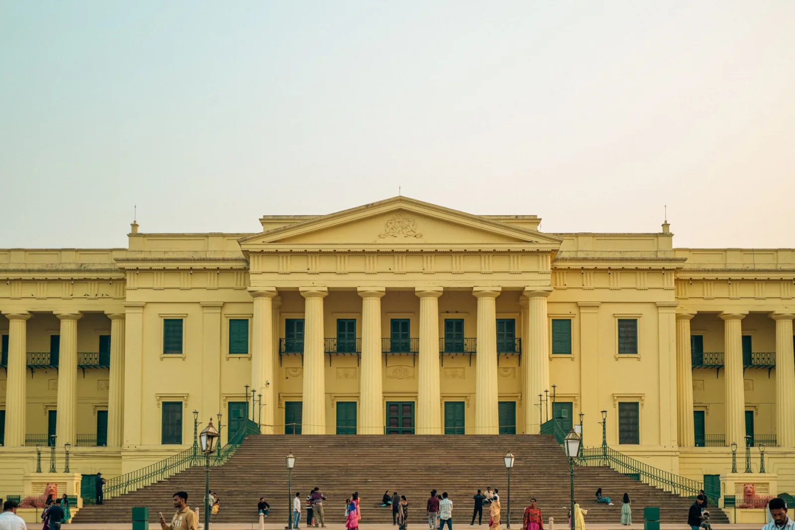 A grand yellow building with columns and people gathering on the steps at sunset.