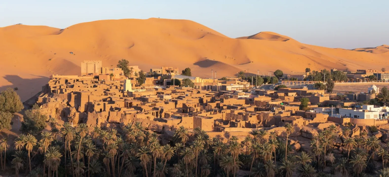 A scenic view of Béchar town in Algeria with desert dunes and palm trees under a clear sky.