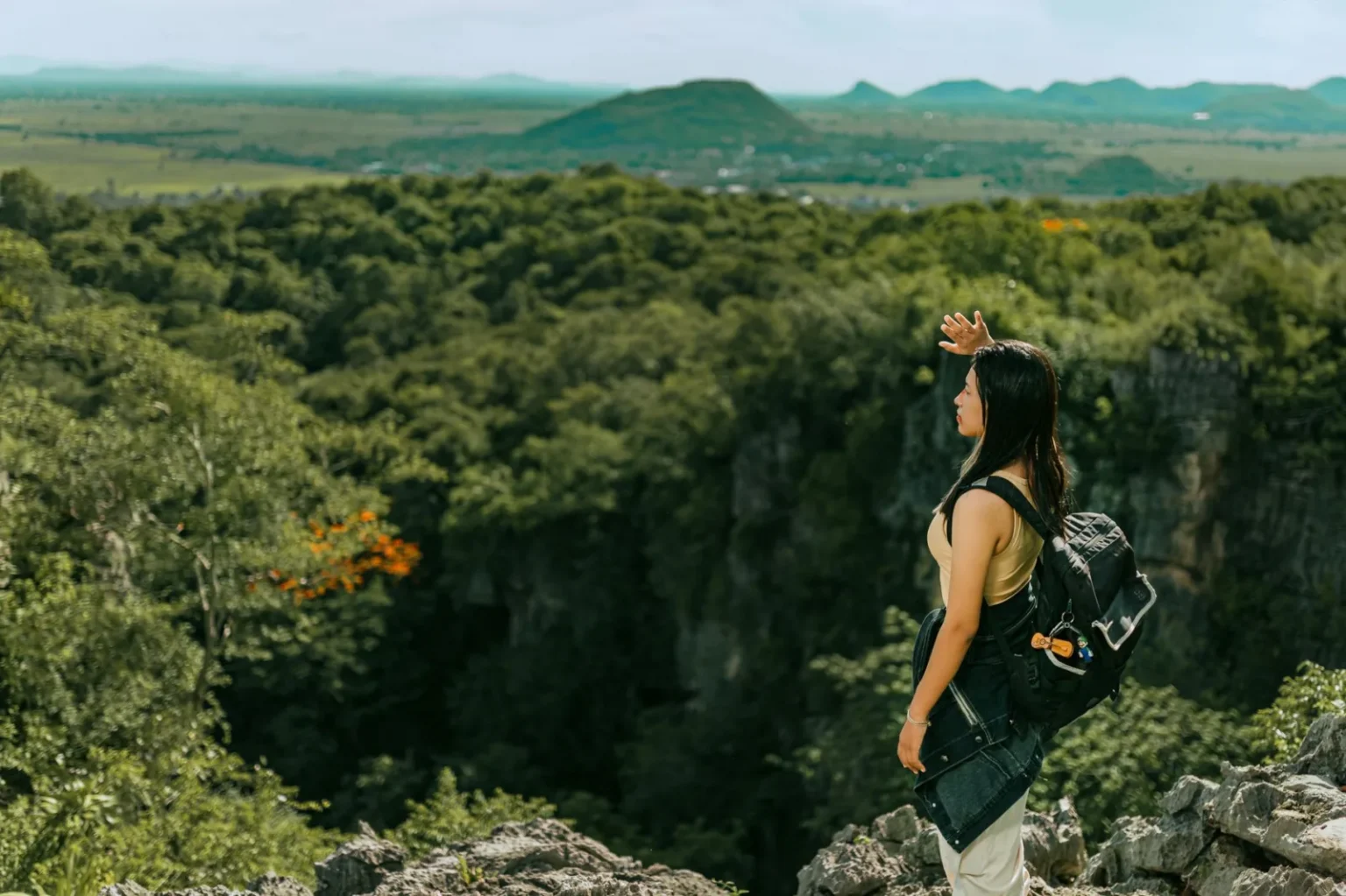 A woman stands on a rocky ledge overlooking lush green forests and distant mountains.