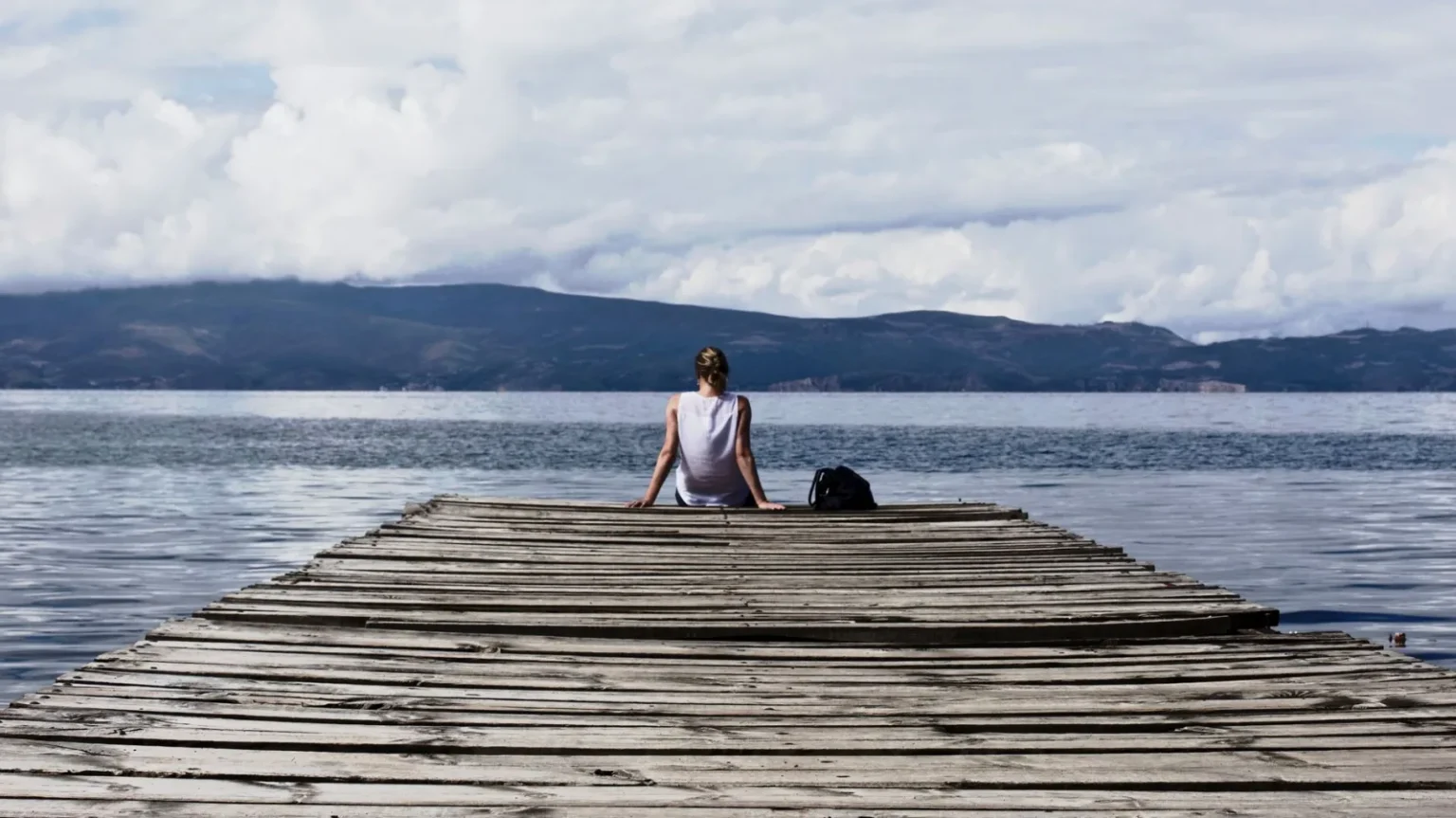 Woman sitting on a wooden pier, enjoying the serene lake view and mountains.