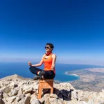 A woman practices yoga on a rocky mountain top with a clear view of the lake below, emphasizing peace and nature.