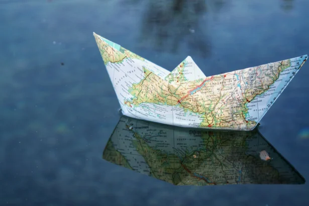 A paper boat folded from a map floats on water, creating a unique reflection.