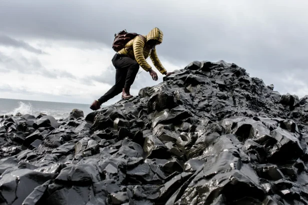 Person in yellow jacket climbs black rocky terrain by the ocean under cloudy skies.