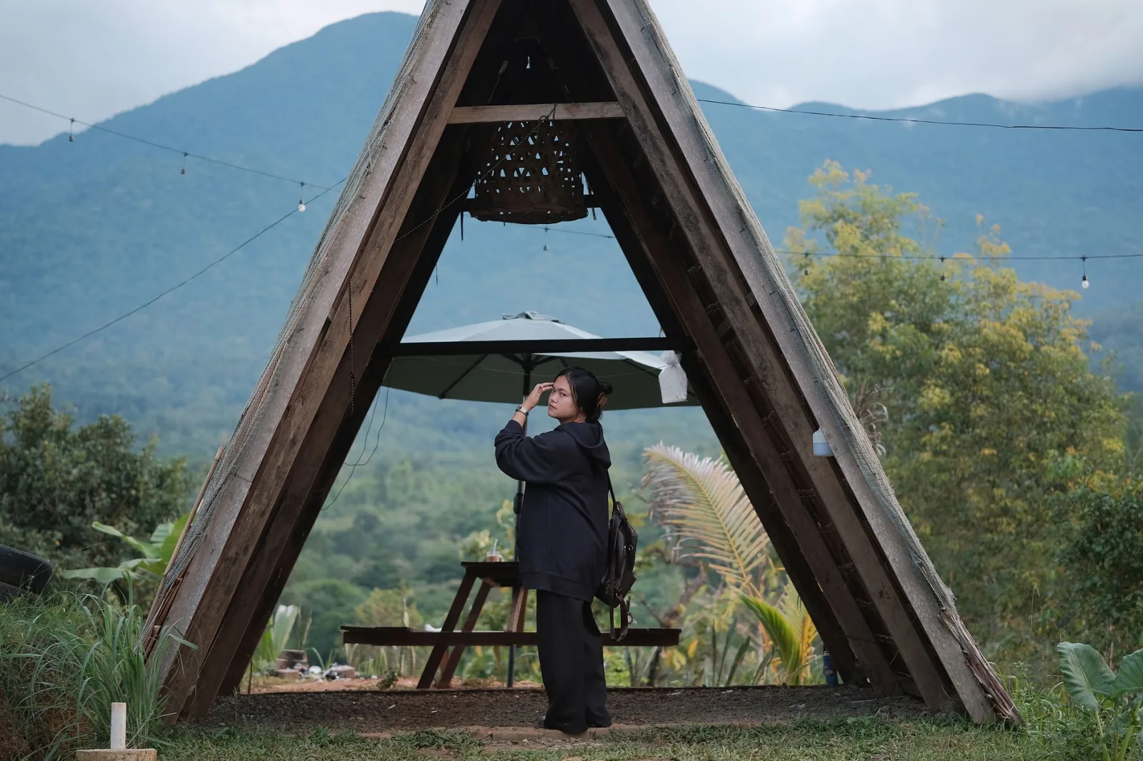 A woman stands under a wooden structure, embraced by Jantho's lush mountains.