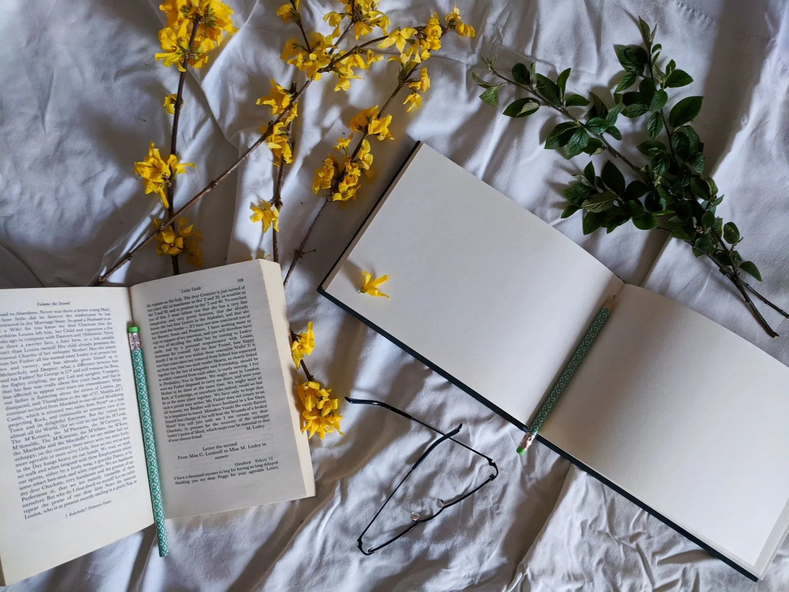 Artistic flat lay of open books, yellow flowers, and pencils on fabric background.