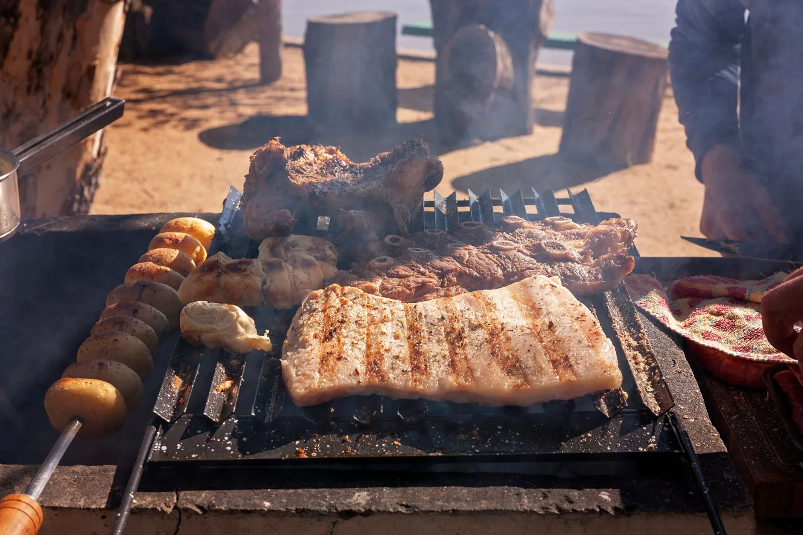 Grilled meats and potatoes on an outdoor barbecue during a sunny day.